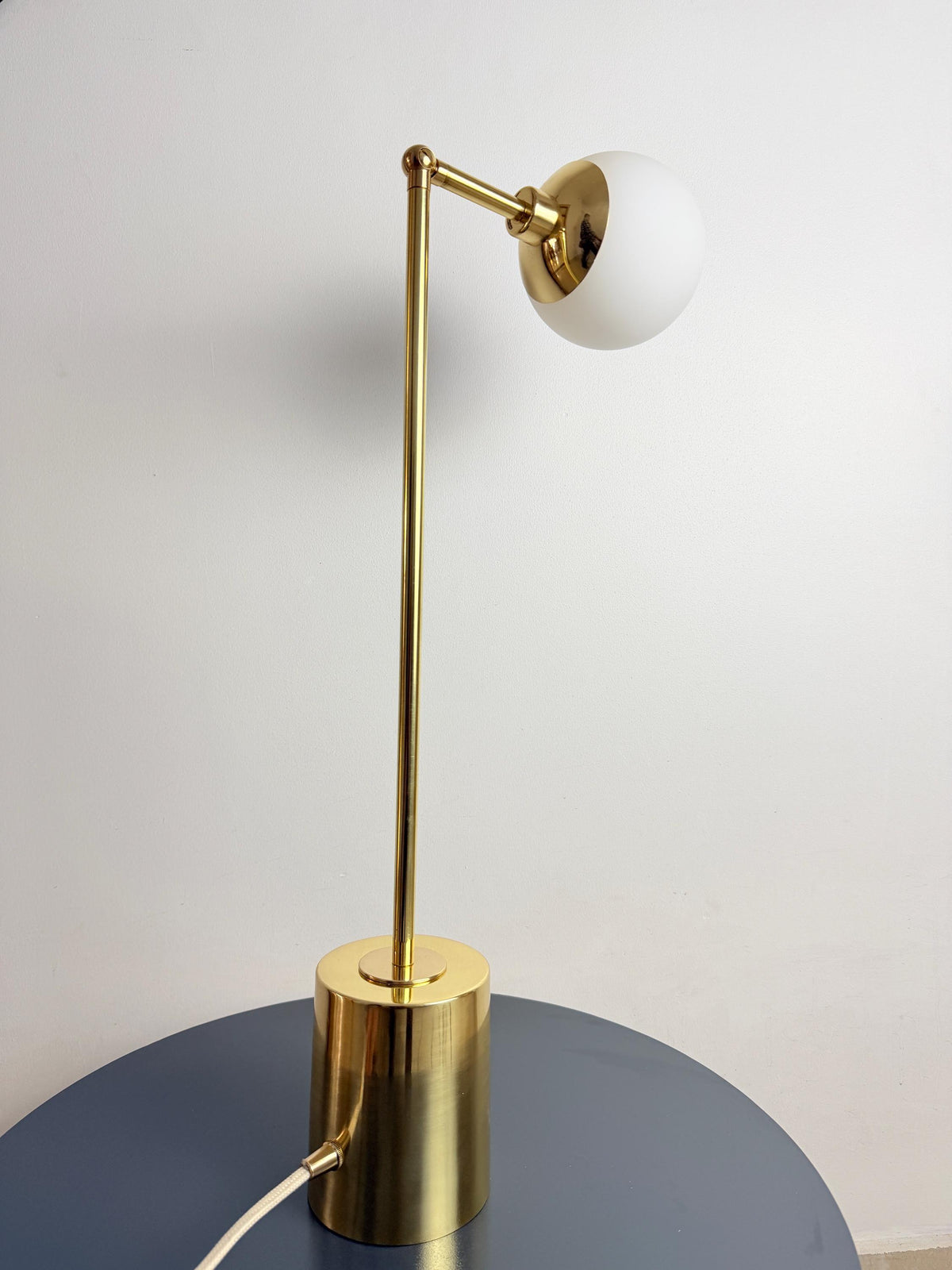 A Clean Contemporary Table Lamp Made with Brass Construction and An Opal White Glass Globe, Featuring an Adjustable Arm Above a Compact Cylindrical Base. the Fixture Has a Restrained Silhouette that Blends Naturally with Modern and Mid Century Interiors, Frost Lights.