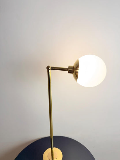 A Sculptural Brass Bedside Lamp with A Smooth Globe Shaped Opal Glass Shade and Adjustable Arm. the Vertical Stem Rises from A Compact Cylindrical Base, Giving This Plug in Table Fixture a Balanced Silhouette with Understated Modern Character, Frost Lights.
